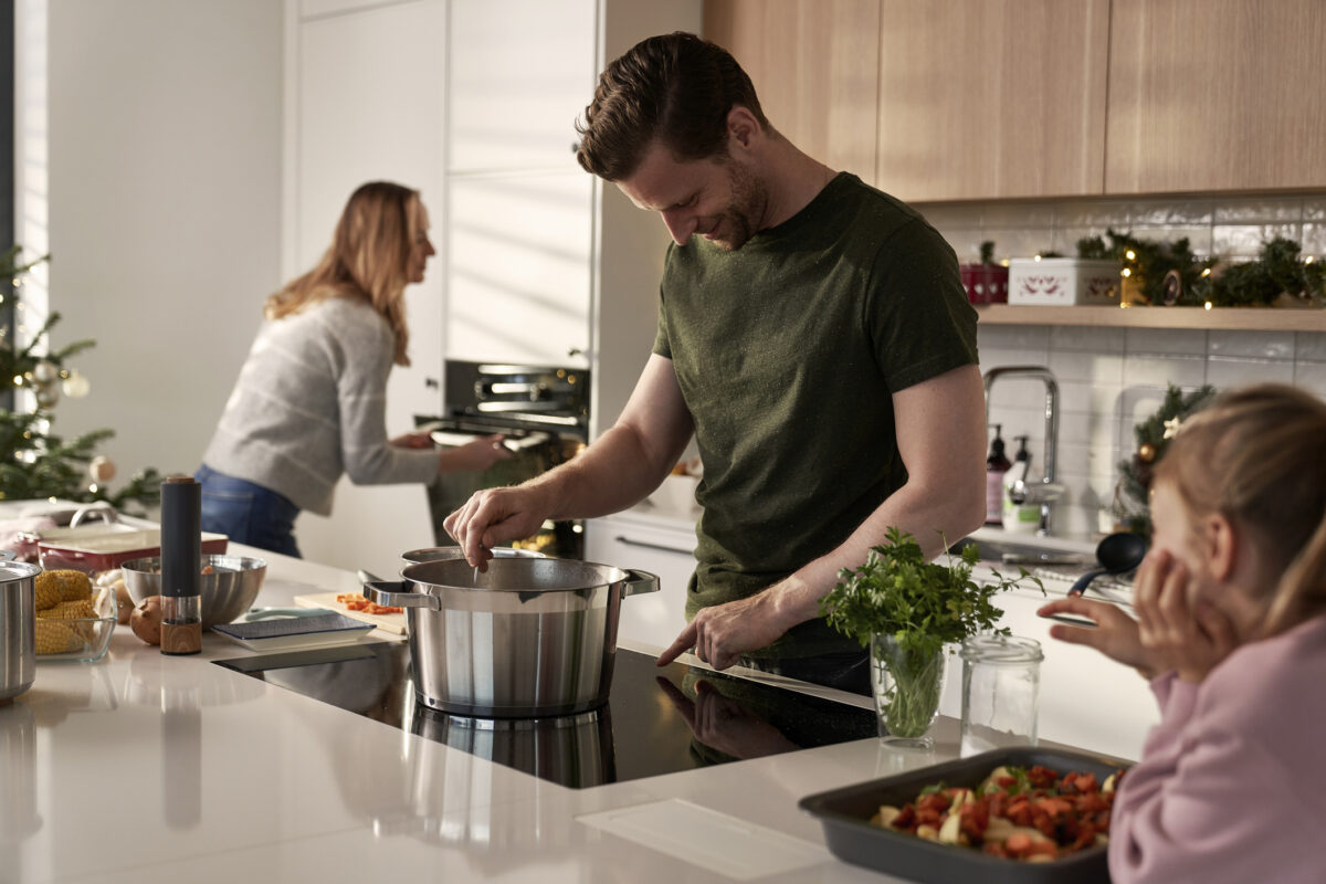 Adult stirring a pot on an induction cooktop in a bright kitchen while another adult prepares food in the background and a child watches from the bench.